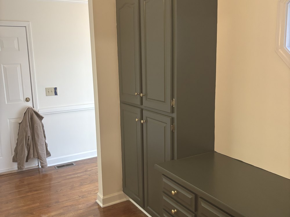A floor-to-ceiling pantry cabinet and a built-in bench finished in dark sage, beside a white-trimmed doorway and a hardwood floor.