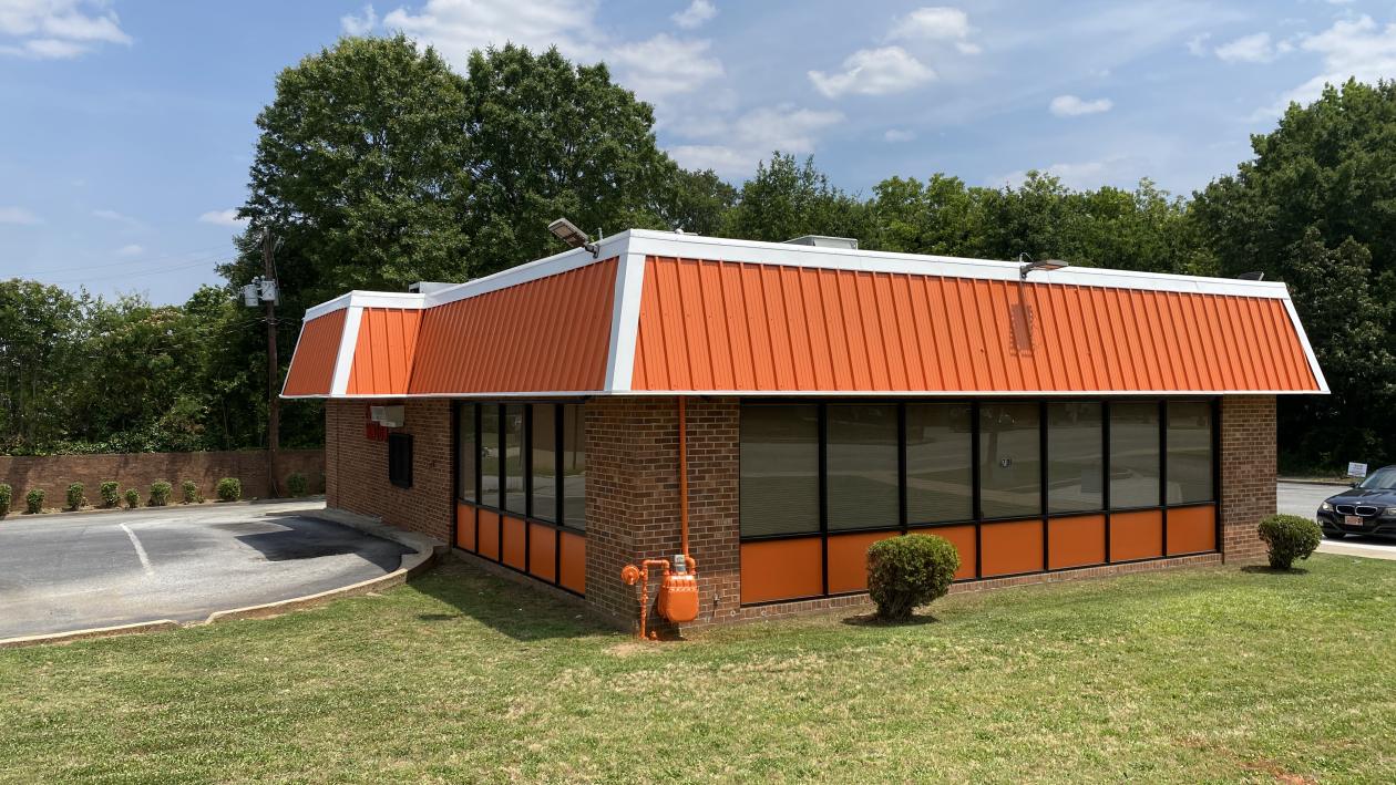 A single-story brick commercial building with an orange standing-seam metal roof and matching orange window panels, seen across a lawn.