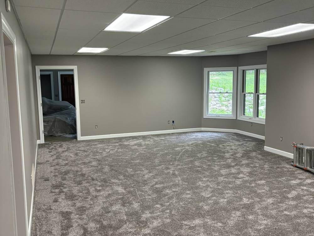 An empty lower-level room with gray painted walls, white trim, a suspended ceiling and new carpet.