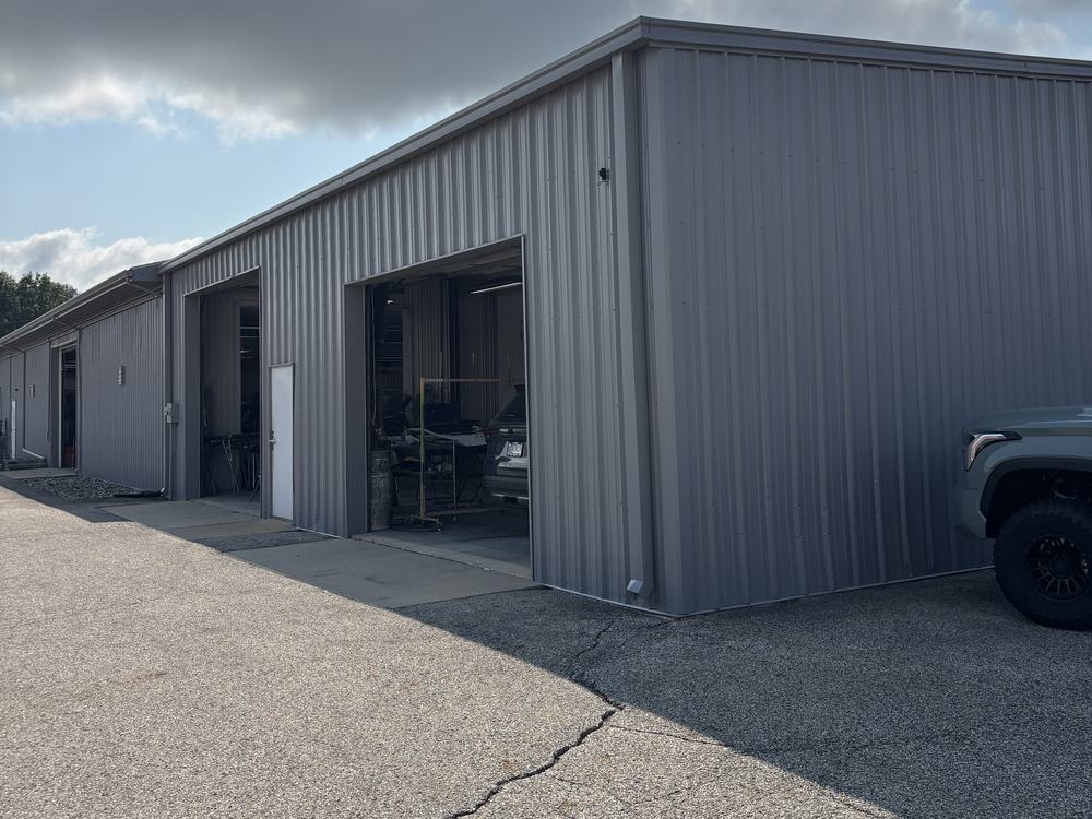 The side of a gray metal commercial building with open roller doors, seen along an asphalt drive under a bright sky.