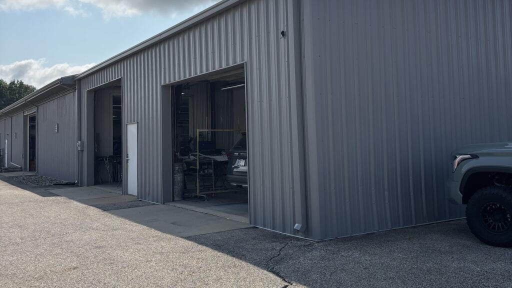 The side of a gray metal commercial building with open roller doors, seen along an asphalt drive under a bright sky.