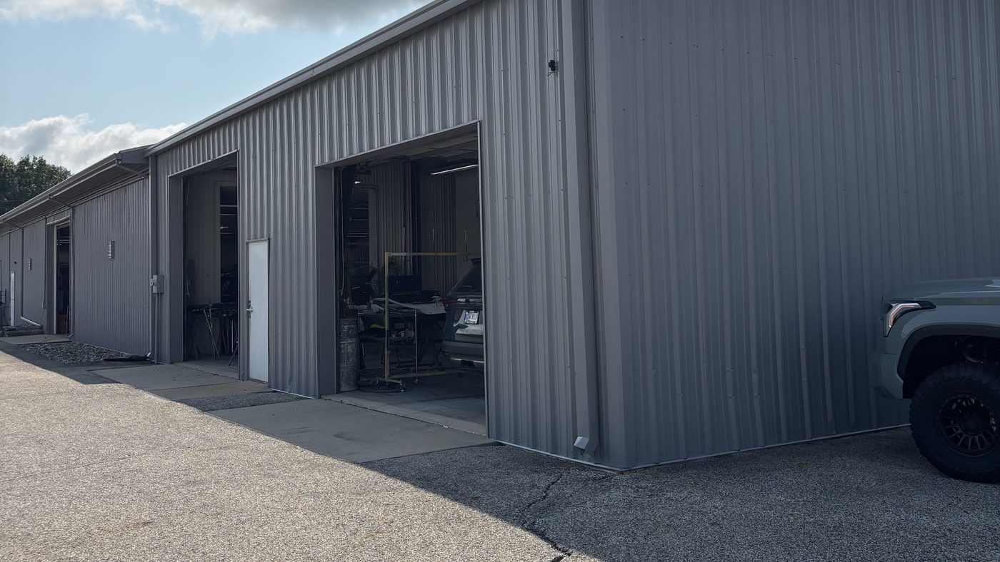 The side of a gray metal commercial building with open roller doors, seen along an asphalt drive under a bright sky.