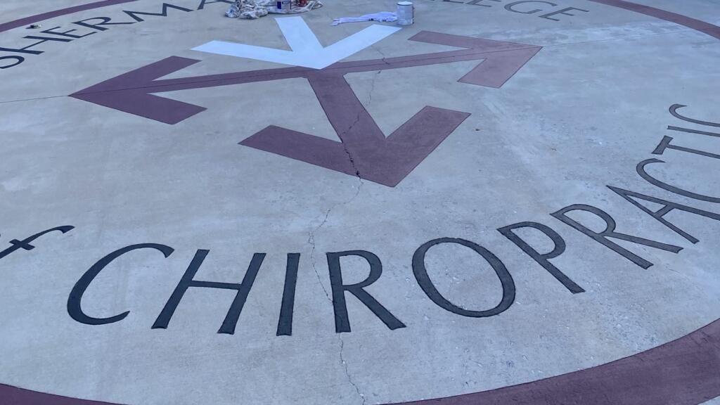 A large seal painted onto a concrete courtyard in maroon and white, reading Sherman College of Chiropractic around a starburst of arrows.