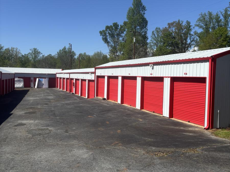 Two rows of self-storage units with red roll-up doors facing each other across a paved drive under a clear sky.