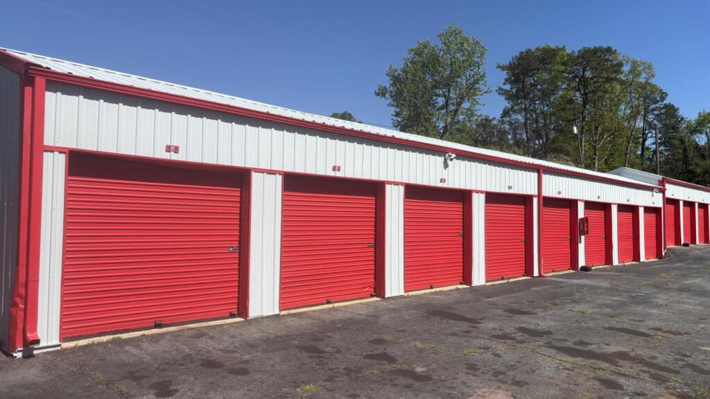A long row of self-storage units with red roll-up doors and red and white metal siding, running away down a paved drive.