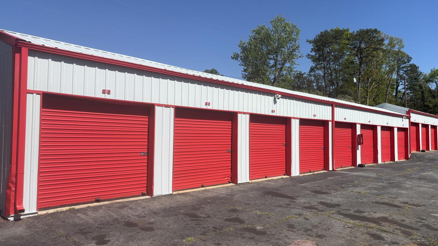 A long row of self-storage units with red roll-up doors and red and white metal siding, running away down a paved drive.