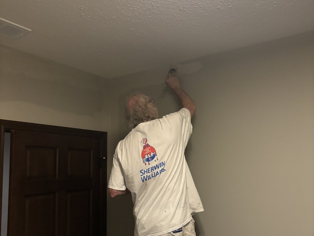 A painter reaching up with a brush to cut in the line where an interior wall meets the ceiling.