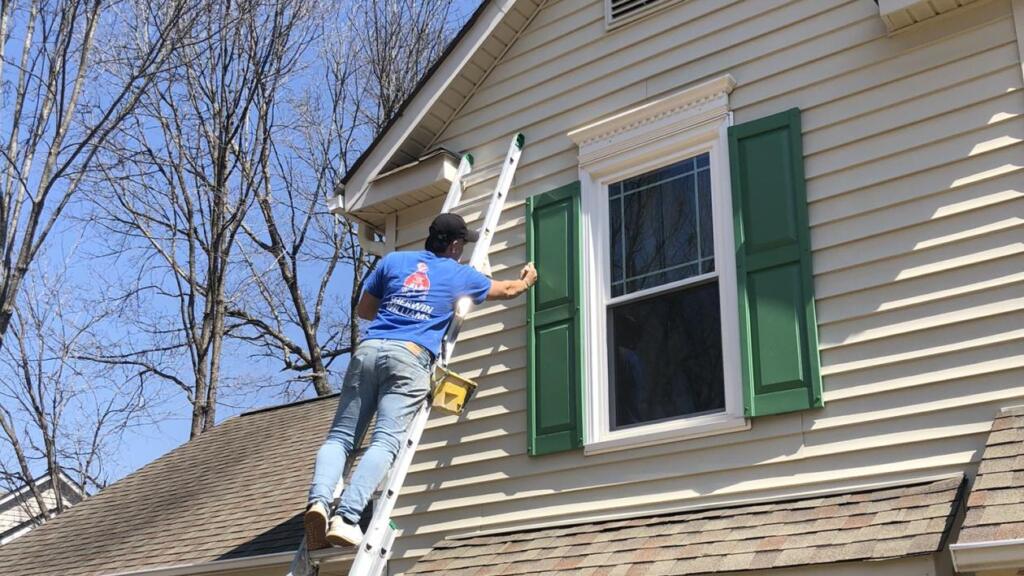 A painter on an extension ladder holding a brush against a green window shutter on the upper story of a cream-sided house, with a paint pail hooked to the ladder rail.