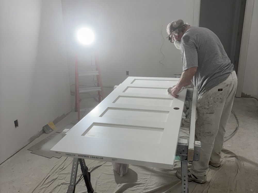 A painter in a respirator spray-finishing a paneled door laid flat on trestles, with drop cloths over the floor and a work light behind.