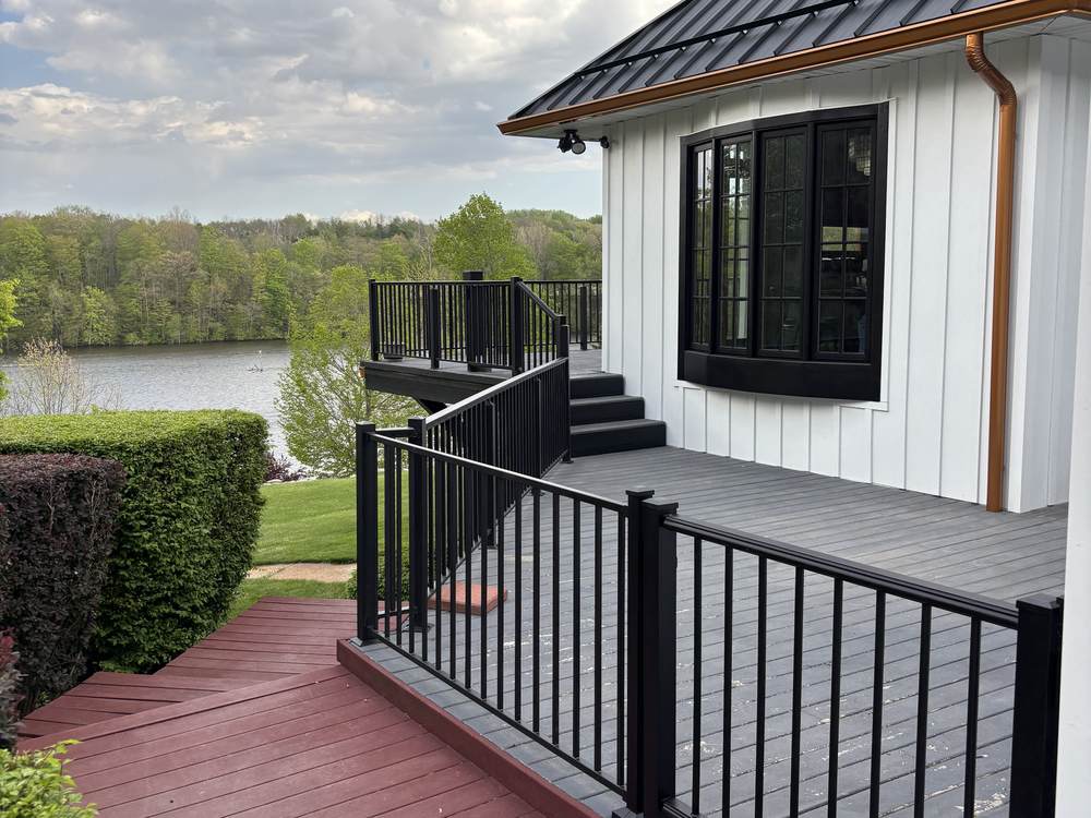 A gray composite deck with black metal railings running beside a white board-and-batten wall, looking out over a lake.