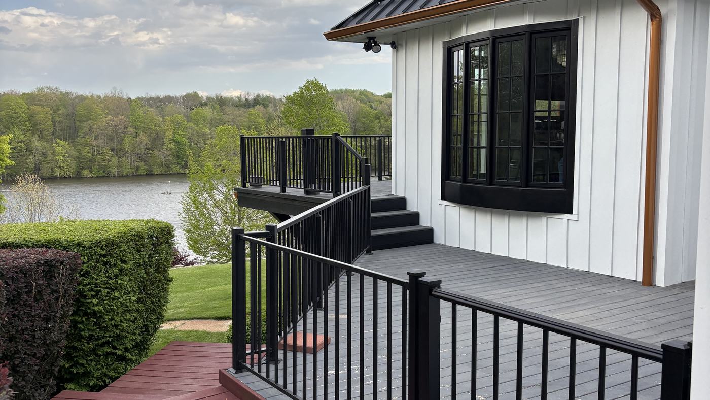A gray composite deck with black metal railings running beside a white board-and-batten wall, looking out over a lake.