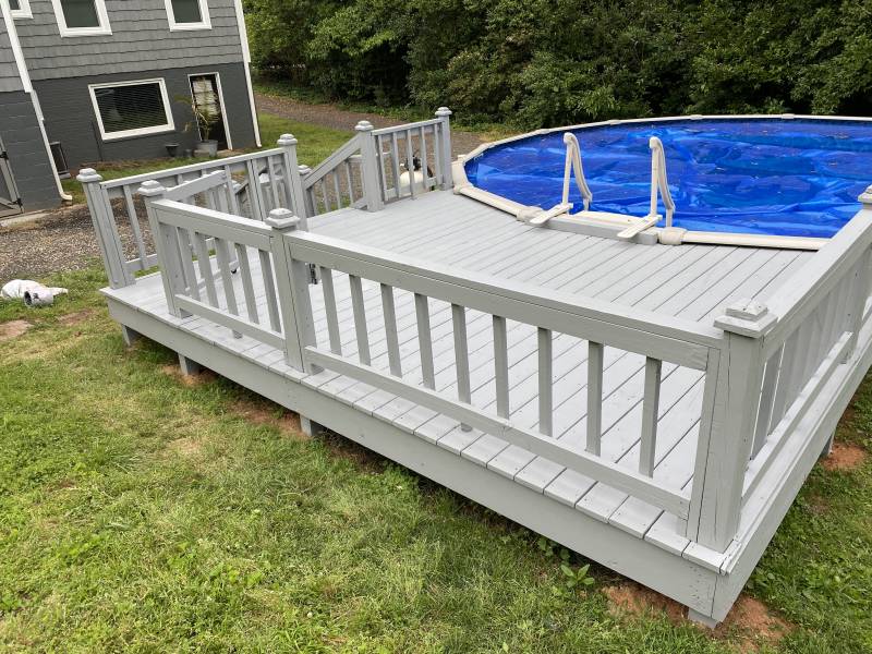 A painted timber deck and railing wrapping an above-ground pool, with steps down to a lawn.