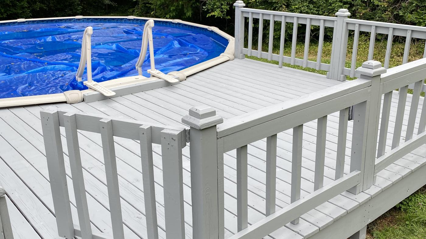 A painted timber pool deck with a white railing and a gate, running alongside an above-ground pool under its cover.