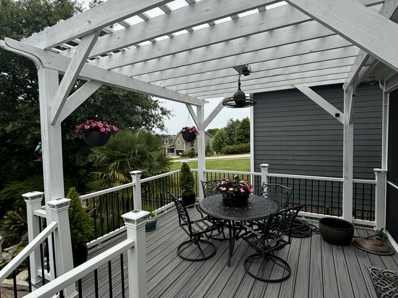 A white-painted pergola over a composite deck with black metal railings, hanging baskets and a table and chairs.