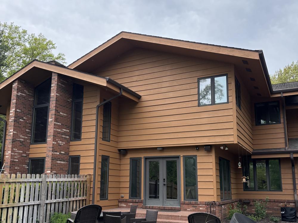The rear of a two-story house with brown wood siding and a brick chimney, above a paved patio with garden furniture.