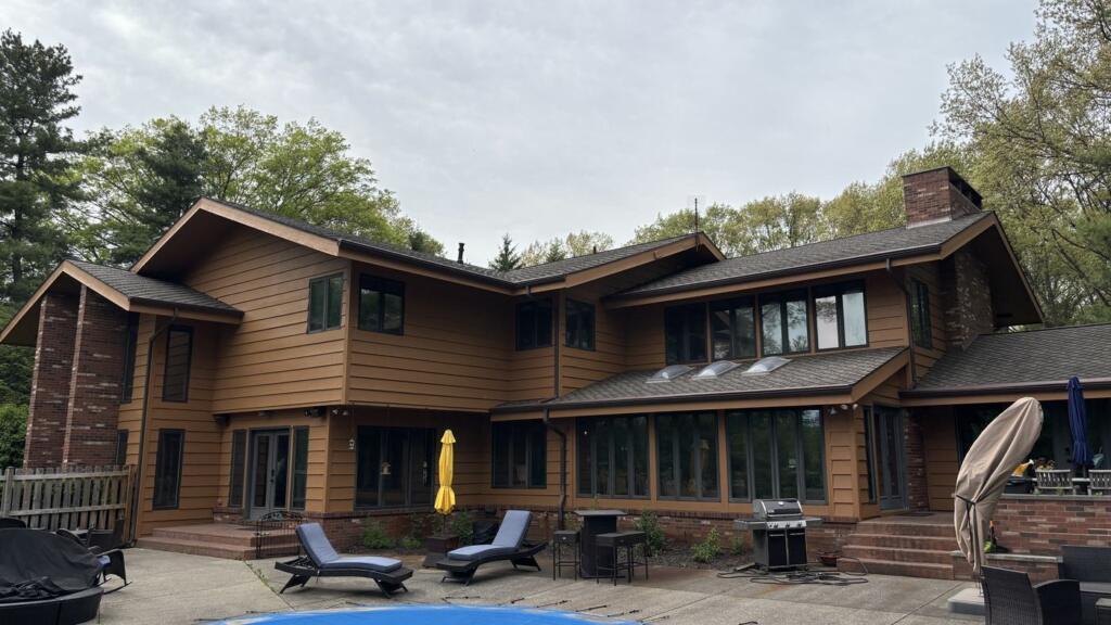 The rear elevation of a two-story house with brown horizontal wood siding and dark window frames, seen across a patio and a covered swimming pool.