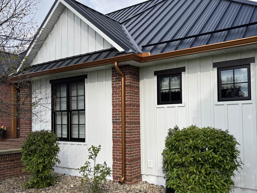 A close view of a painted gable end where white board-and-batten siding meets a dark metal roof, with a copper downspout running down a red brick column.