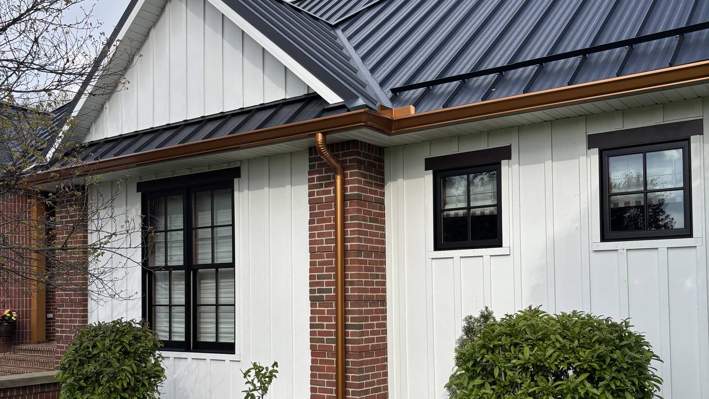A close view of a painted gable end where white board-and-batten siding meets a dark metal roof, with a copper downspout running down a red brick column.