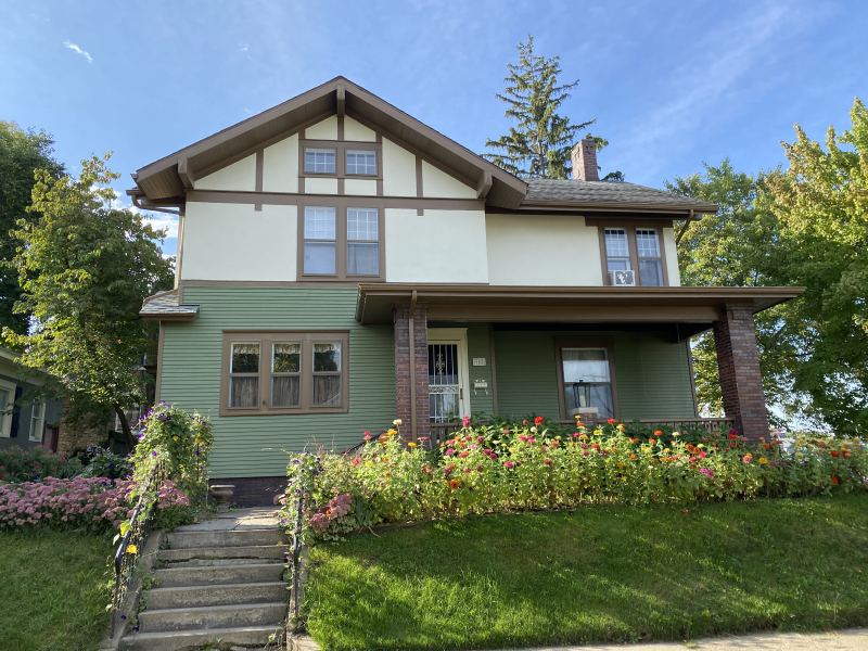The front of a sage-green craftsman house with cream gable stucco, dark brown trim and a brick-columned porch, seen from the sidewalk across a flower border.