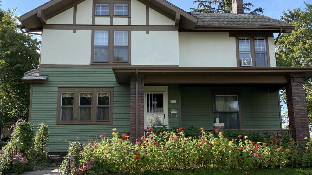 A two-story craftsman house painted sage green with cream gable stucco and dark brown trim, above a brick-columned porch and a border of zinnias.