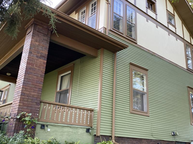 The side of a craftsman house in sage green with dark brown window casings, a red brick porch pier and a turned timber rail.