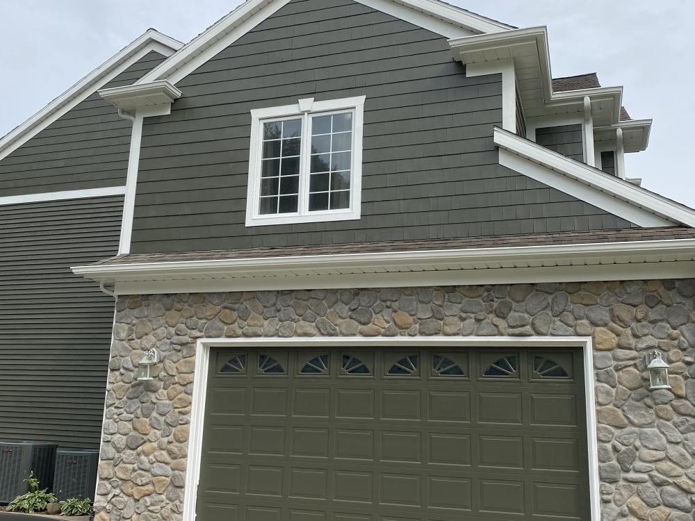 An olive-green gable and garage door above a river-rock stone base, with white trim and painted eave brackets.