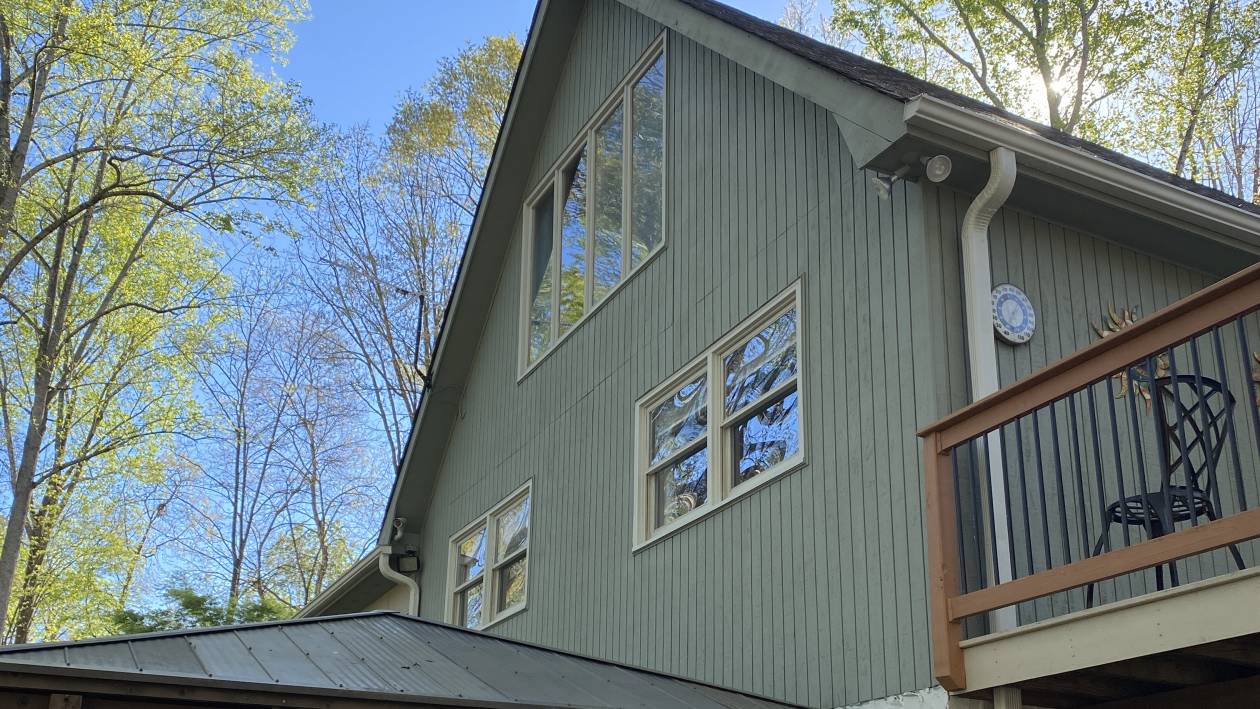 The gable end of a house clad in sage-green board-and-batten siding, with white window trim and a timber porch roof below.