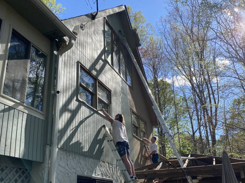 Two painters working from ladders on the board-and-batten gable of a house among trees, the lower half already recoated.
