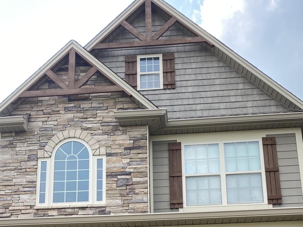 A gable end faced in gray shake siding and stacked stone, with timber brackets, an arched window and dark brown shutters.