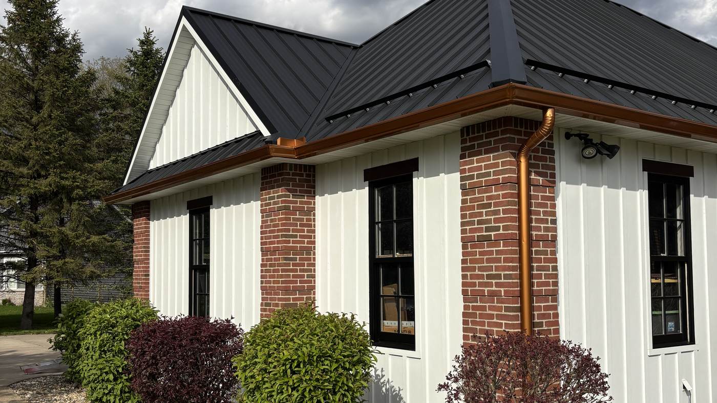 A single-story house with white board-and-batten siding, red brick columns and a dark standing-seam metal roof, with copper gutters running along the eave.