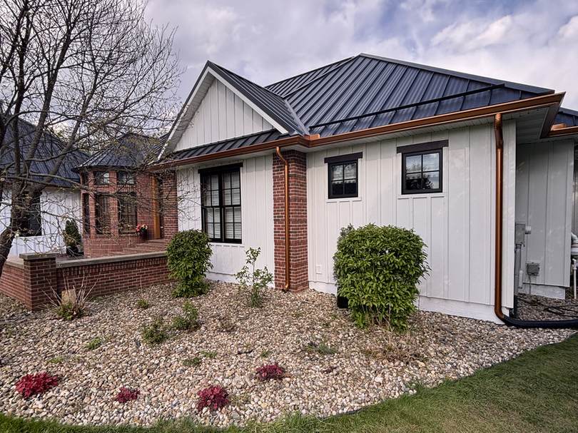 The front corner of a house with white board-and-batten siding, a red brick column and a dark metal roof, with copper downspouts above a gravel planting bed.