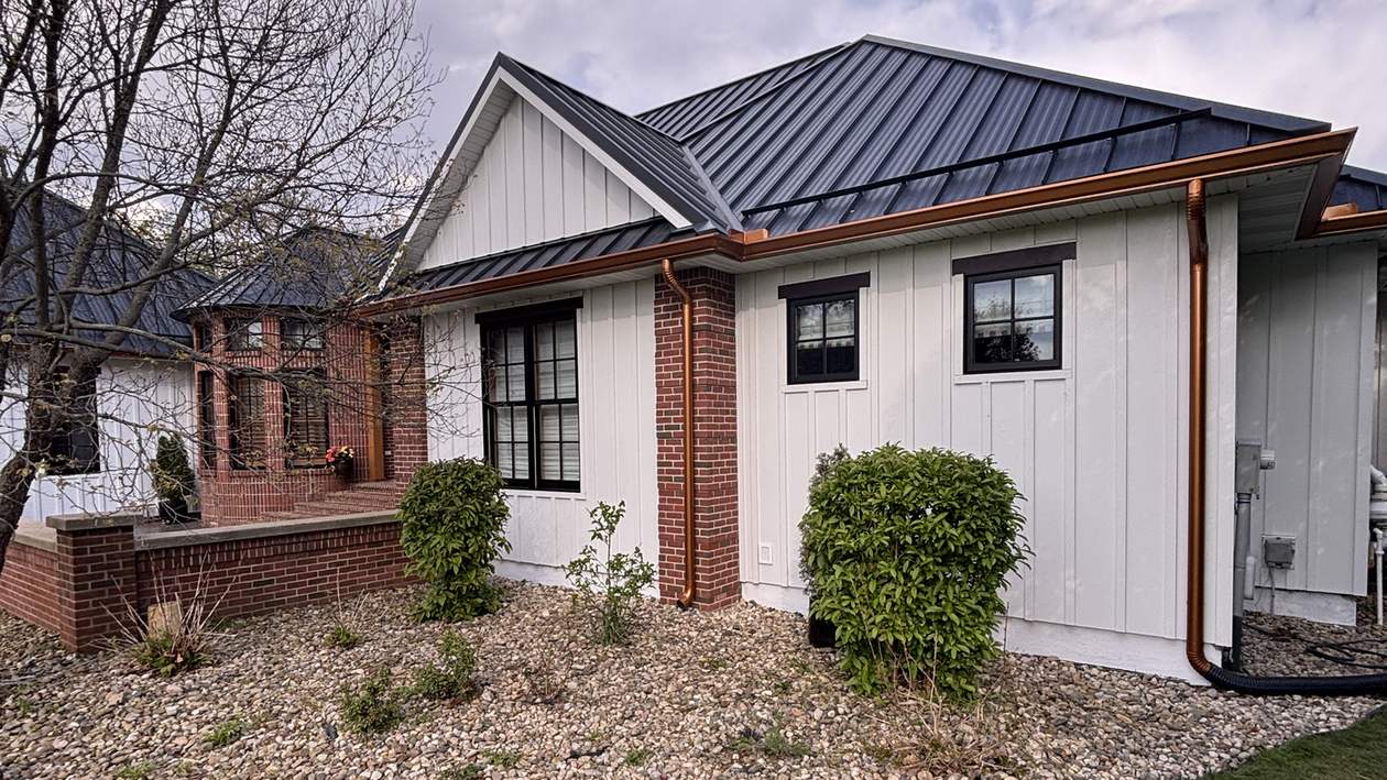 The front corner of a house with white board-and-batten siding, a red brick column and a dark metal roof, with copper downspouts above a gravel planting bed.