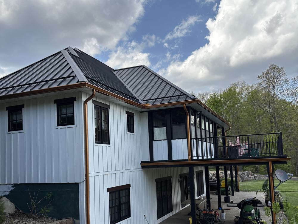 A white board-and-batten house with a dark metal hipped roof and copper gutters, with a screened porch and a railed deck above a walkout lower floor.
