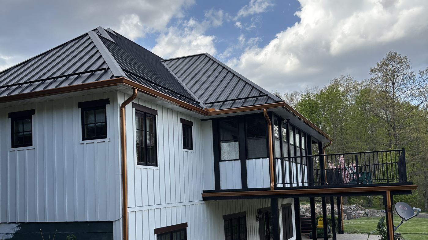 A white board-and-batten house with a dark metal hipped roof and copper gutters, with a screened porch and a railed deck above a walkout lower floor.