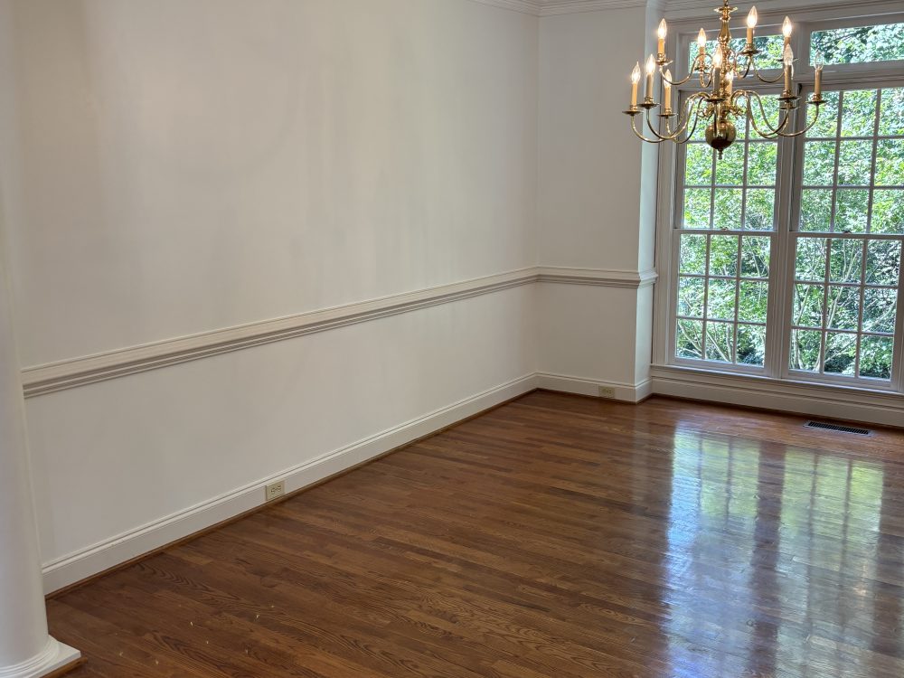 An empty dining room with pale painted walls, a white chair rail and crown molding, a chandelier and a hardwood floor.