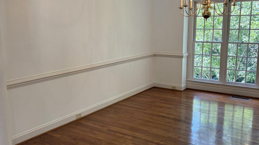 An empty dining room with pale painted walls, a white chair rail and crown molding, a brass chandelier and a hardwood floor.