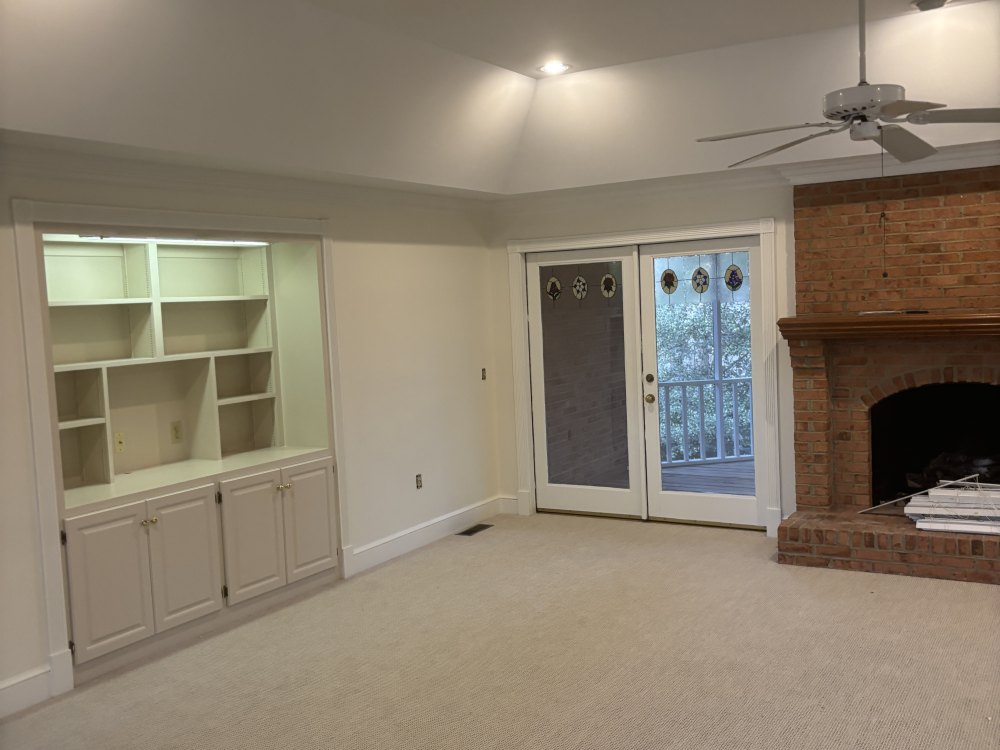 An empty living room with freshly painted walls, white built-in shelving beside glazed doors, and a red brick fireplace.