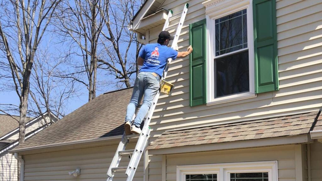 A painter near the top of an extension ladder set against a cream-sided house, working on the green shutters at an upstairs window.